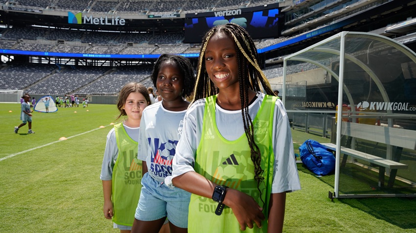 three girls smile at the camera. they are standing in a sort of diagonal line -- at the front is a black girl with long thin braids about 13 years old wearing a bright green pinny over a light blue U.S. Soccer Foundation jersey. the girl in the middle is black and has short black hair. the girl at the back is white and shorter with brown hair. they are also wearing light blue U.S. Soccer Foundation jerseys.